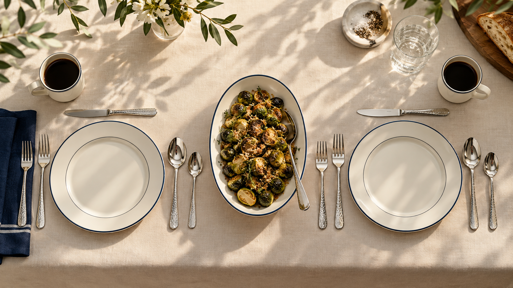 Sunlit Chefcrest table for two with five-piece hammered cutlery settings, ivory and navy plates, and roasted Brussels sprouts